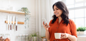 Young woman drinking coffee and looking at her smart phone while standing in her kitchen